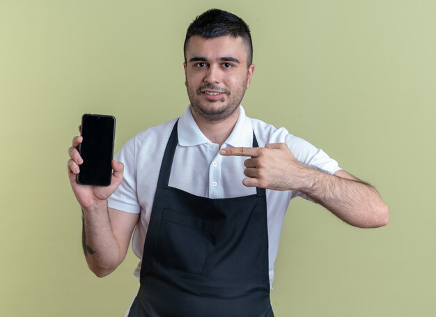 Barber man in apron showing smartphone pointing with index finger at it smiling confident