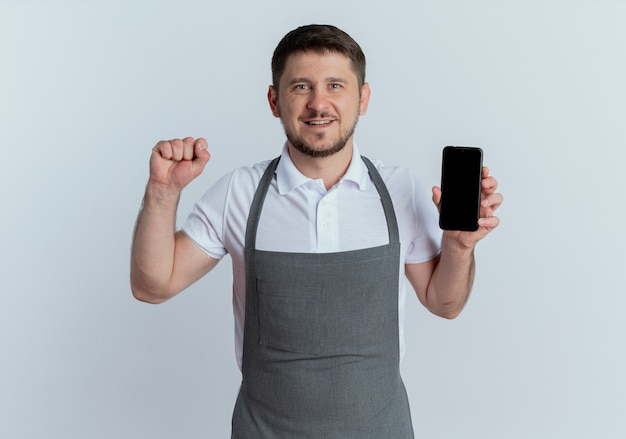 Barber man in apron showing smartphone raising fist happy and positive looking at camera standing over white background