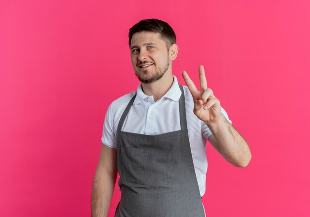 Barber man in apron smiling cheerfully looking at camera showing victory sign standing over pink background