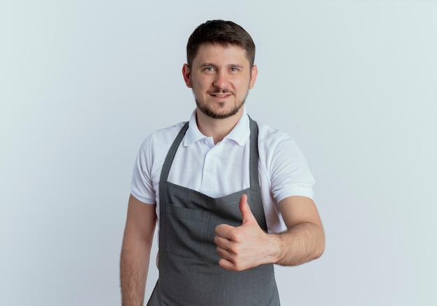 Uomo barbiere in grembiule sorridente fiducioso che mostra i pollici in su in piedi sul muro bianco