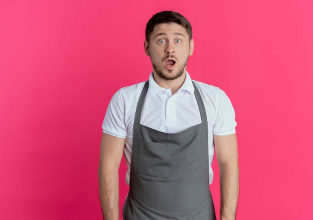 Barber man in apron surprised and amazed standing over pink wall