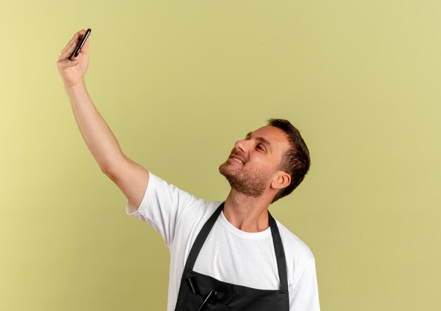 Парикмахер в фартуке, делающий селфи с помощью своего смартфона, весело улыбаясь, стоя над светлой стеной