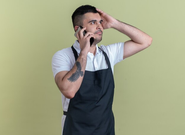 Barber man in apron talking on mobile phone looking aside smiling confident standing over green background