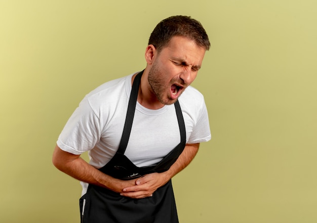 Free photo barber man in apron touching his belly having pain looking unwell standing over light wall