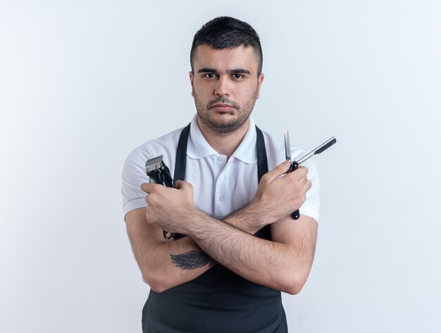 Парикмахер в фартуке с парикмахерскими инструментами, глядя в камеру с серьезным уверенным выражением лица, стоящим на белом фоне
