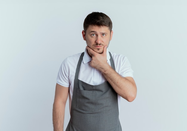 Uomo barbiere in grembiule con la mano sul mento pensando in piedi sul muro bianco