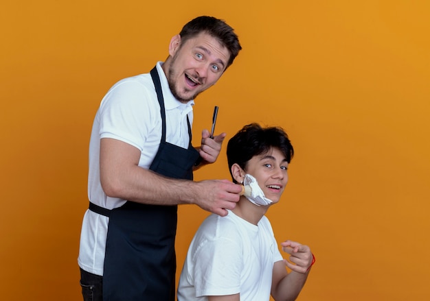 barber man in apron  with happy face pointing with finger putting shaving foam with shaving brush on face of satisfied client over orange wall