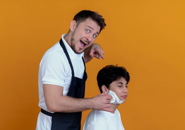 barber man in apron  with happy face putting shaving foam with shaving brush on face of satisfied client over orange wall