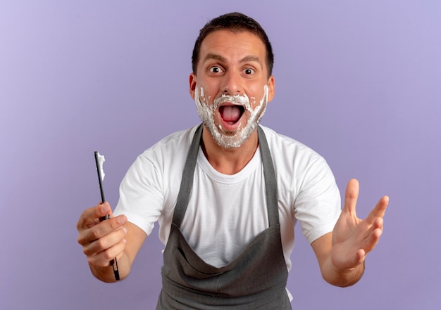 Barber man in apron with shaving foam on his face holding razor looking to the front surprised and happy standing over purple wall