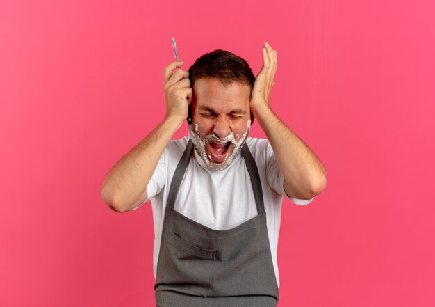 Barber man in apron with shaving foam on his face, holding razor with hands on his head shouting with aggressive expression standing over pink wall