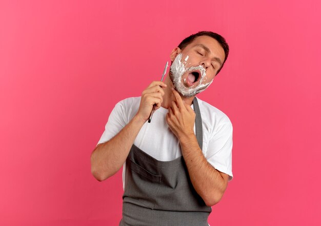 Парикмахер в фартуке с пеной для бритья на лице бреет бороду бритвой, стоя над розовой стеной