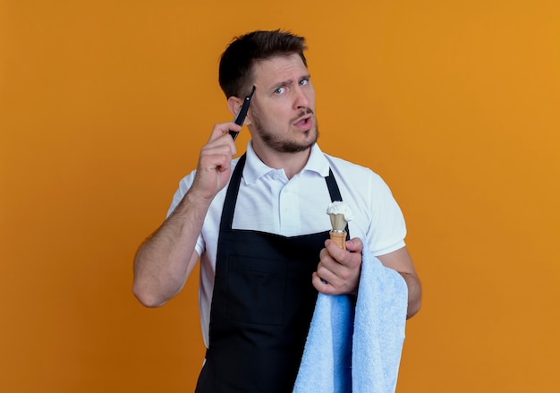 Barber man in apron with towel on his hand holding shaving brush with foam and razor looking at camera puzzled standing over orange background