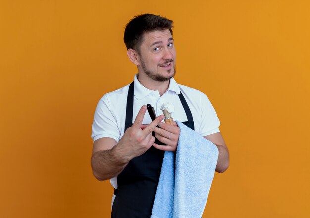 Barber man in apron with towel on his hand holding shaving brush with foam and razor looking at camera smiling confident showing number two standing over orange background