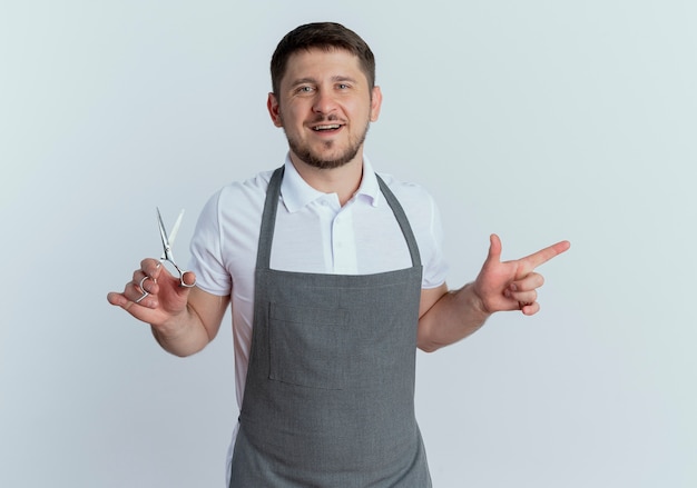 Бесплатное фото Парикмахер в фартуке держит ножницы, указывая указательным пальцем в сторону, улыбаясь, стоя над белой стеной
