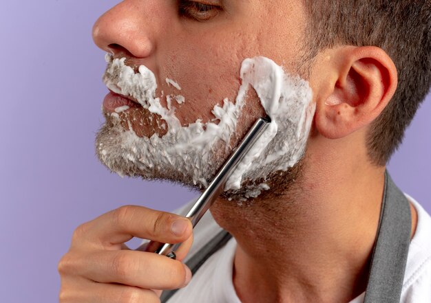 Barber man with shaving foam on his face shaving himself using razor over purple wall