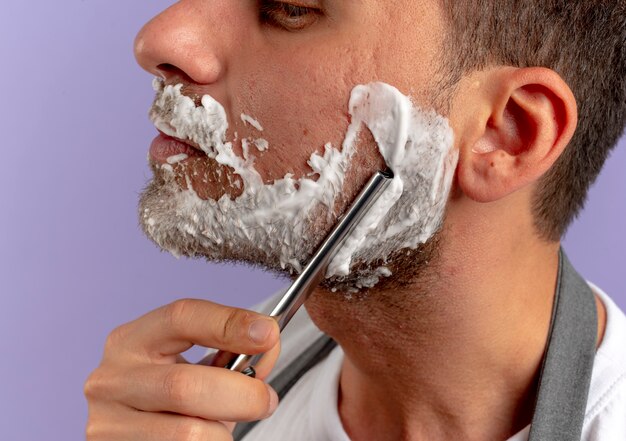 보라색 벽 위에 면도기를 사용하여 자신을 면도하는 그의 얼굴에 거품을 면도하는 이발사 남자