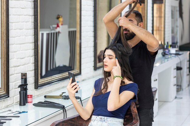 Парикмахер растягивает волосы девушки39, а девушка смотрит в свой телефон Фото высокого качества