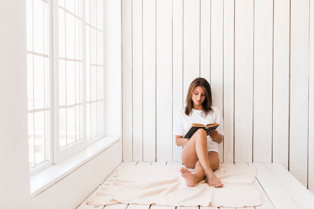 窓の近くの毛布で読んでいる裸足の女性