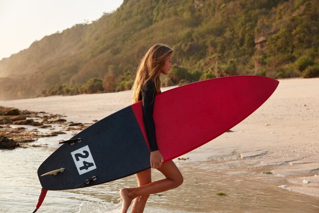 Barefoot woman stands sideways, holds surfboard, enjoys free time for surfing, poses in ocean near shore, against rock