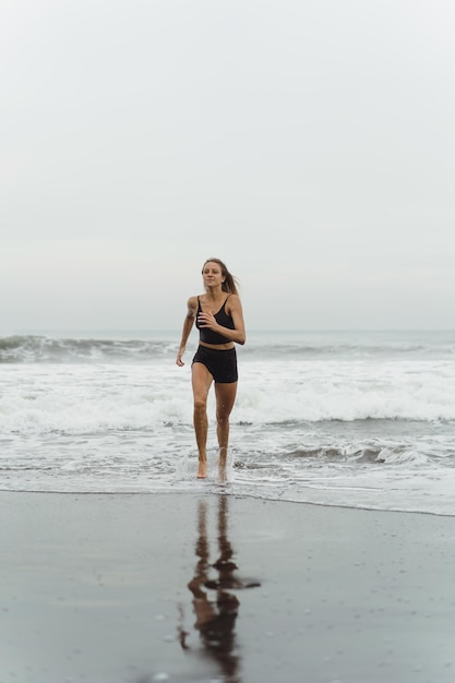 Free photo a barefoot young woman with a slender body runs on the sea surf by the water pool to keep fit and burn fat. beach background with blue sky. women's fitness, jogging, sports activities on a summer fami