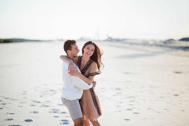 白い砂の上に明るい刺繍の服を抱きしめる柔らかい裸足のカップル