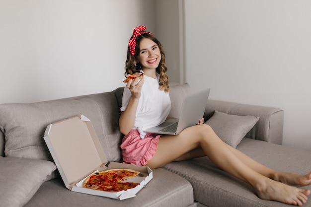 Barefooted elegant girl watching movie and eating pizza. Indoor shot of smiling female freelancer using laptop and enjoying italian food.