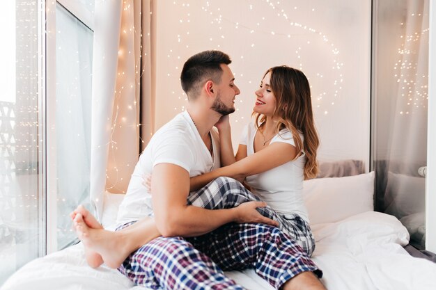 Barefooted lady in cozy pajama sitting on boyfriend's knees. Glad female model looking into husband's eyes.