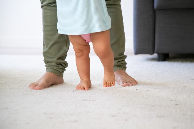 Barefooted legs of baby trying to walk in front of mom. Kid making first steps with moms support. Cropped shot. Parenthood and childhood concept