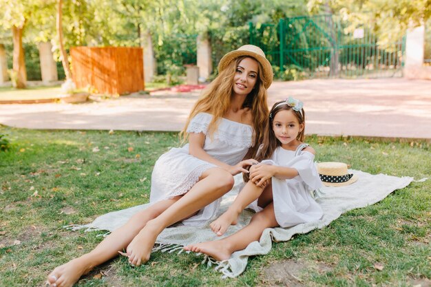 Barefooted long-haired girl relaxing on blanket with little sister and sunbathing in sunny day. Outdoor portrait of smiling young woman chilling on the grass with cute daughter in elegant dress.