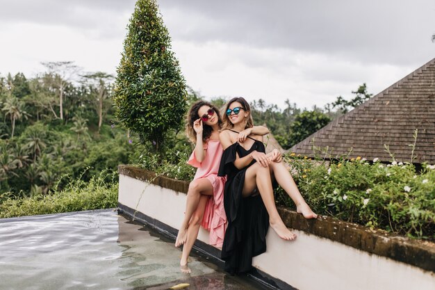 Barefooted woman in black dress posing with sister near swimming pool. Laughing female models in sunglasses relaxing at Bali in vacation.