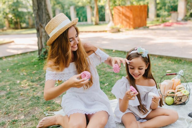 Barefooted woman in hat with white ribbon sitting on blanket near daughter and eating cookies smiling. Outdoor portrait of happy family joking and fooling around during picnic.