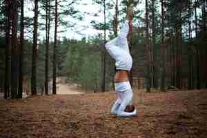 Free photo barefooted young male in white clothes doing variation of salamba shirshasana yoga stance on ground in forest, crossing legs. outdoor shot of advanced yogi training in woods, balancing on hands