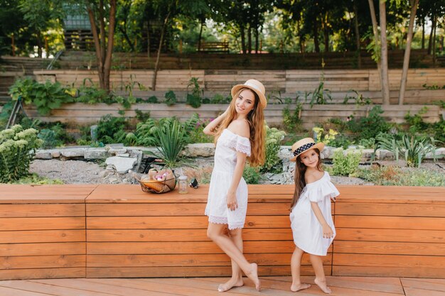 Barefooted young woman and little girl standing back to back in front of flowerbed. Outdoor full-length portrait of stylish mother and daughter wearing similar outfit in summer park.