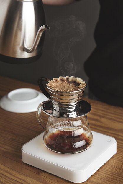 블랙 스웨트 샷의 바리 스타는 여과 된 커피 / 실버 찻 주전자를 흰색 단순한 무게의 아름다운 투명한 크롬 드립 커피 메이커에 준비합니다. 카페 숍의 두꺼운 나무 테이블에있는 모든 것. 증기