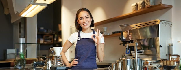 カウンターのバリスタは ⁇ OK ⁇ のサインを表示し ⁇ 笑顔のアジア人女の子がカフェで働き ⁇ 青いエプロンを着て ⁇ おすすめします ⁇ 