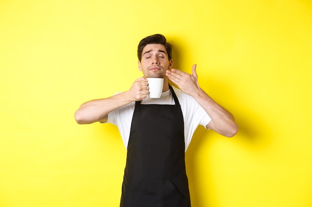 Barista enjoying smell of coffee in mug, standing pleased with eyes closed, wearing black apron.