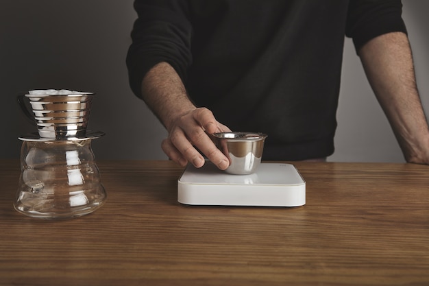 Free photo barista holds stainless silver cup with roasted ground coffee above white simple weights. drip coffee maker for filtered coffee near.