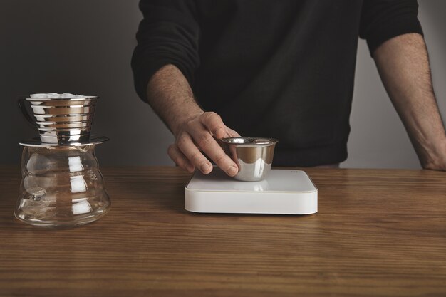 barista holds stainless silver cup with roasted ground coffee above white simple weights. Drip coffee maker for filtered coffee near.