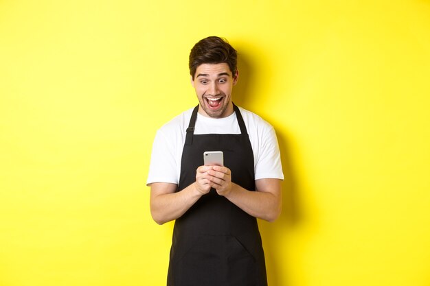 Barista looking surprised as reading message on mobile phone standing in black apron against yellow ...