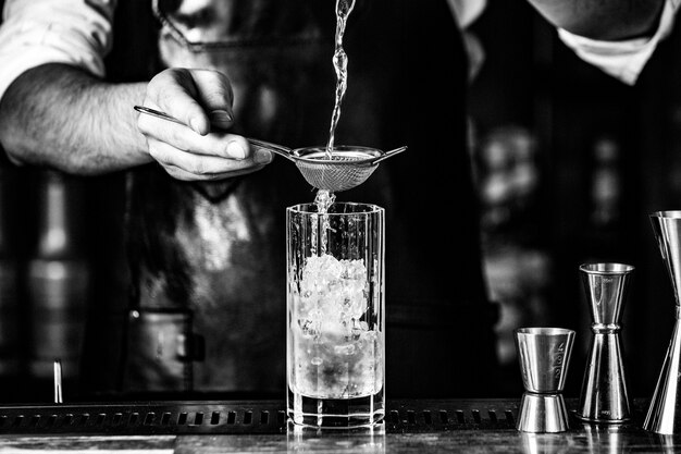 Barista putting alcohol into a cocktail glass with syrup and ice cubes.