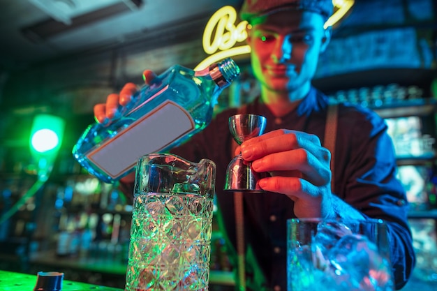 Barman finishes preparation of alcoholic cocktail with shaker in multicolored neon light