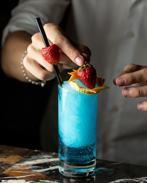 Bartender prepares blue cocktail garnished with orange zest and strawberries