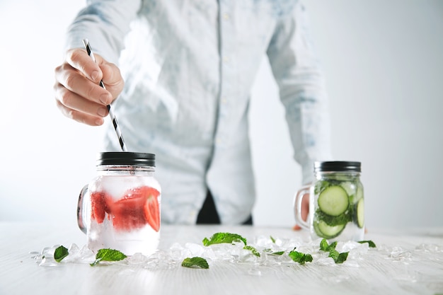 Bartender puts striped drinking straw in jar with fresh cold homemade lemonade made from ice, strawberry, crashed melted ice and cucumber mint lemonade in other jar on back.