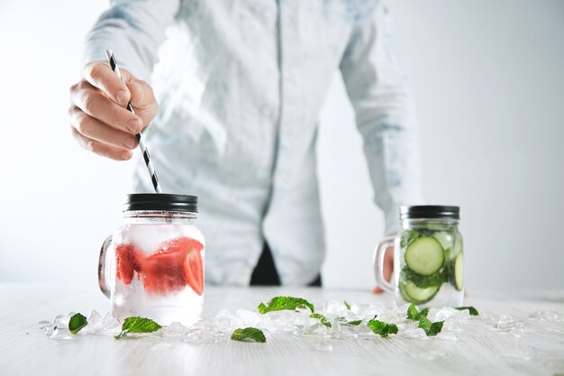 Bartender puts striped drinking straw in jar with fresh cold homemade lemonade made from ice, strawberry, crashed melted ice and cucumber mint lemonade in other jar on back.