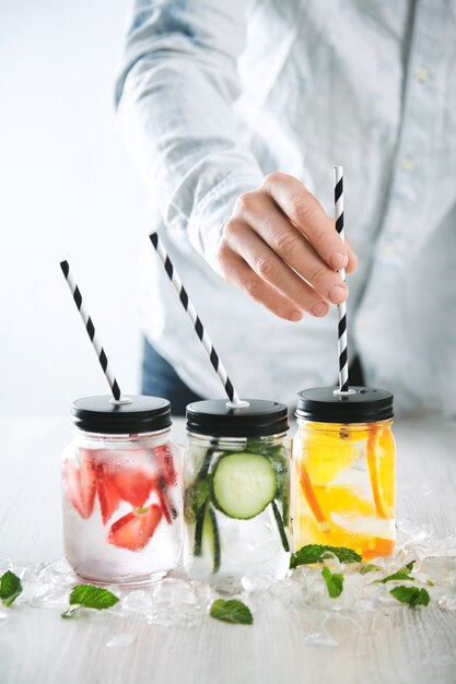 Bartender는 얼음, 딸기, 오렌지, 오이 및 민트로 만든 신선한 차가운 수제 레모네이드와 함께 병에 줄무늬 음료수 빨대를 넣습니다.