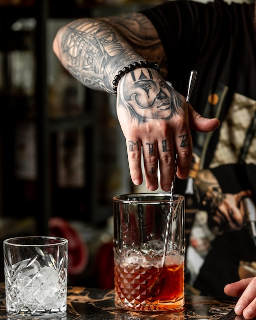 Бармен с татуировками делает красный коктейль с виски.