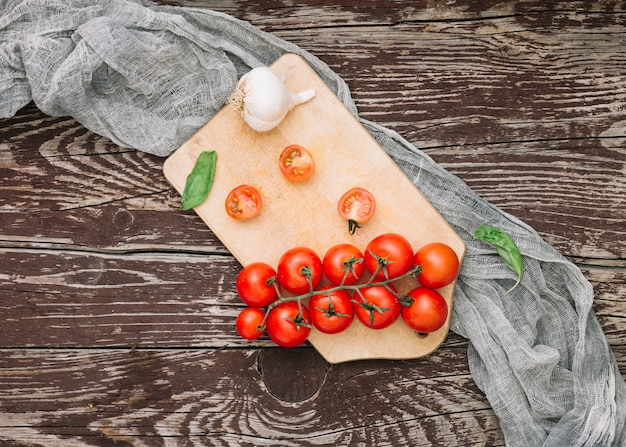 Basil; cherry tomatoes and garlic cloves on chopping board with grey cloth over the wooden textured backdrop