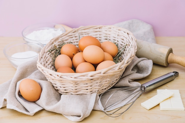 Basket full of whole eggs; flour; sugar; white chocolate; whisks and rolling pin on wooden desk