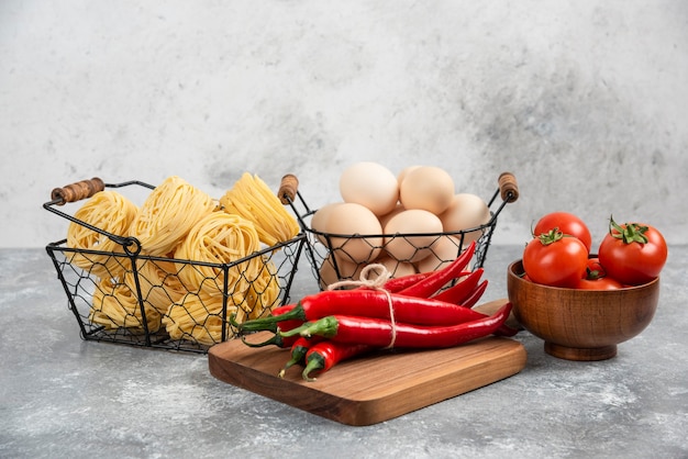 Basket of raw noodles, tomatoes, chili peppers and eggs on marble surface.