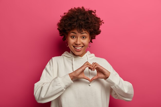 Be my valentine. Pleasant looking curly woman expresses love, makes heart gesture, has romantic feelings